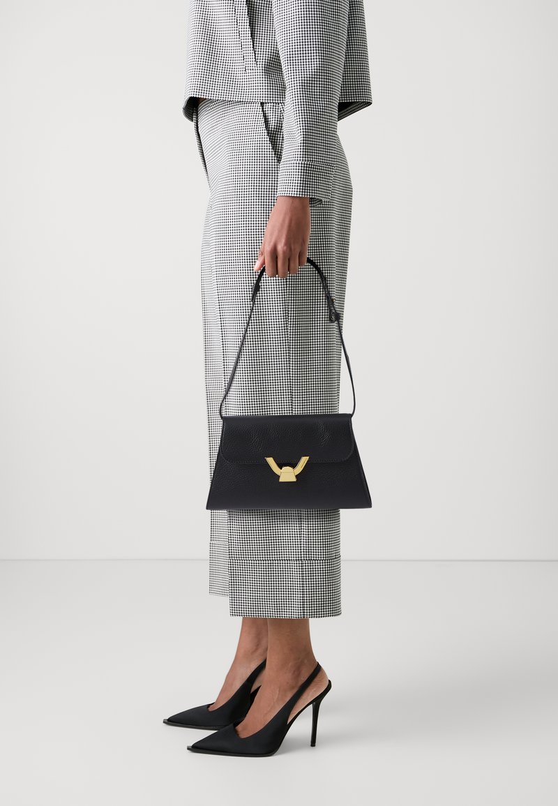Borsa a tracolla in pelle nera con una chiusura dorata, forma rettangolare e manico corto, posizionata accanto a un completo a scacchi e tacchi a punta.