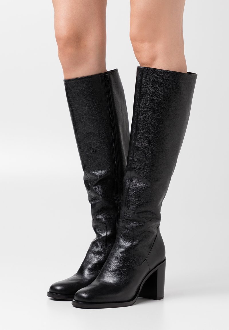 Bottes en cuir noir au-dessus du genou avec un bout arrondi, un talon épais et une fermeture éclair latérale. La texture est lisse avec de légers détails de plis.