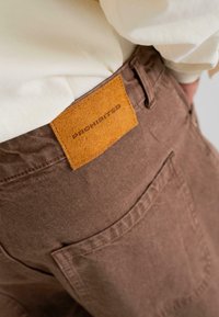 Pruunist korduroost valmistatud püksid, mille taga on suede oranž silt, millel on kiri "KEELATUD". Omab taskut ja silmatorkavaid õmbluse detaile.