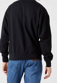 Felpa nera con una vestibilità rilassata, polsini e orlo a coste e una texture in cotone morbido. Indossata con jeans in denim blu con tasche posteriori.