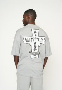 Camiseta oversized de color gris claro con un diseño gráfico en negro de una cruz y el texto "MULTIPLY APPAREL EST 2015" en la parte posterior.