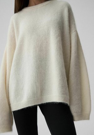 Kobieta nosząca luźny, miękki, kremowy sweter z długimi rękawami, w połączeniu z czarnymi spodniami na neutralnym tle.