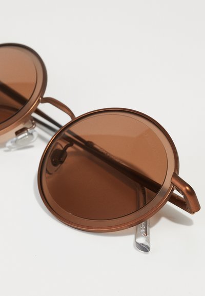 Brown circular sunglasses with a metallic frame, featuring tinted lenses and clear temple tips for comfort. Details include a minimalist design.