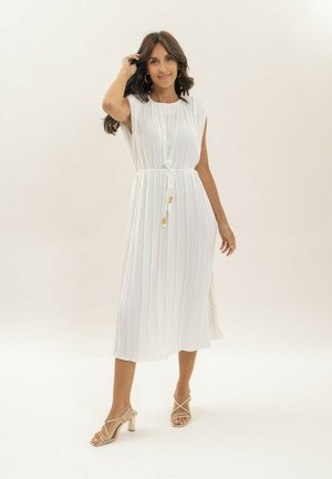 Femme portant une robe midi blanche plissée sans manches avec ceinture à la taille et sandales à talons beiges, debout devant un fond uni.