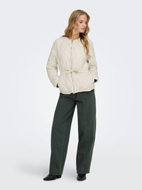 Femme aux longs cheveux blonds portant une veste matelassée beige clair, un pantalon large foncé et des bottines noires, debout les mains dans les poches.