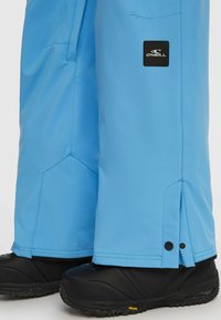 Blauwe sneeuwbroeken met een gladde afwerking, voorzien van een zijrits en een logo-patch. Gedragen over zwarte sneeuwlaarzen met gestructureerde zolen.
