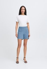 Shorts de mezclilla en azul claro con un corte clásico, que cuentan con dos bolsillos frontales y un dobladillo sin terminar. Combinados con una camiseta blanca ajustada y sandalias negras.