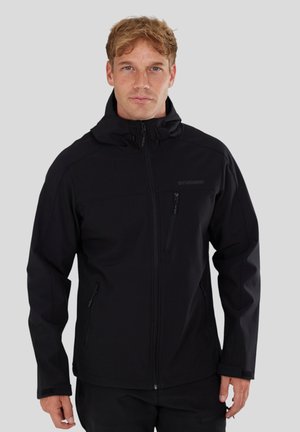 Man wearing a black zip-up jacket with hood, standing against a plain light gray background, facing forward.