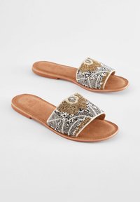 Perlenbesetzte Riemchensandalen mit einem geschwungenen Riemen, ausgestattet mit goldenen, silbernen und schwarzen Perlen auf einem hellbraunen Wildledersohle und -fußbett.