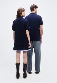 Δύο άτομα στέκονται πλάτη προς το μέρος μας, φορούν μπλε μπλουζάκια πόλο· το ένα φοράει μαύρη φούστα με μαύρες μπότες, το άλλο μπλε τζιν με μαύρα παπούτσια.