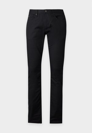 Homme portant un t-shirt noir Emporio Armani, un jean noir et des baskets noires, debout avec une main dans la poche sur un fond blanc uni.