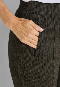 Zwart en bruin broek met houndstooth-patroon met een zijzak, waar een hand gedeeltelijk in is gestoken. De stof lijkt textuur te hebben en is op maat gemaakt.
