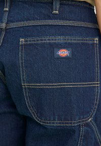 Temno modri traper hlače z zadnjim žepom, rumenim šivom in oznako Dickies na desni strani.