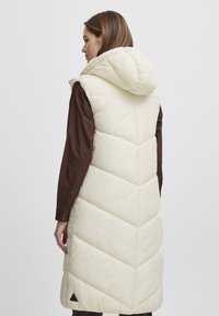 Gilet long en duvet crème avec un design en chevrons, capuche ajustable, et un accent triangulaire noir à l'ourlet. Chemise marron en dessous.