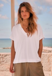 Haut blanc à manches courtes et col en V en tissu doux, associé à un pantalon ample marron clair, sur fond de paysage côtier.