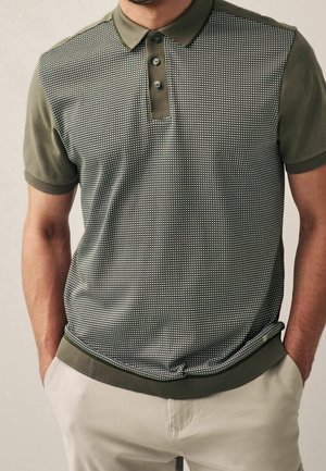 Mand iført olivengrøn polo med mønstret frontpanel og beige bukser, hænderne i lommerne.