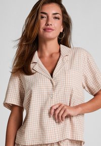 Korte mouw, knoopsluiting overhemd in lichtbeige met wit geruit patroon, voorzien van een zak en zachte textuur. Model weergegeven op een neutrale achtergrond.