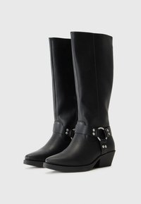 Botas de cuero negras hasta la rodilla con punta afilada, tacón bajo y apilado, y una correa decorativa con acentos de metal plateado.