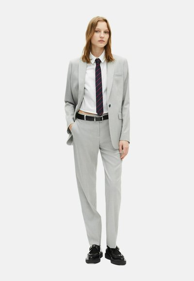 Completo su misura grigio chiaro con rever affilati, taschino singolo, abbinato a una camicia bianca e una cravatta a righe navy. Cintura nera e mocassini completano il look.