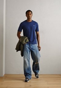 Marineblå t-shirt med lille logo, lyseblå slidte jeans og mørke sko. Modellen holder en grøn jakke over den ene arm.
