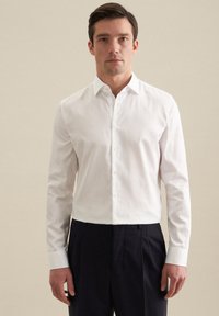 Biała koszula z długim rękawem, zapinana na guziki, o klasycznym kołnierzyku, wykonana z gładkiego materiału, w zestawieniu z ciemnymi spodniami. Prosty design, bez widocznych wzorów.