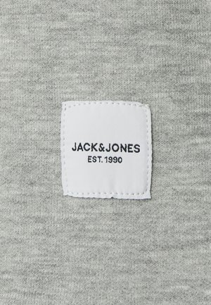 Harmaa kangas, jossa on neulottu tekstuuri ja valkoinen kudottu etiketti, jossa lukee "JACK&JONES EST. 1990" mustalla tekstillä.