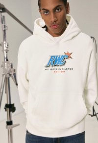 Fehér kapucnis pulóver, elülső zsebbel, "RWC" grafikai logóval és "WE MOVE IN SILENCE EST.1991" felirattal kék és narancssárga színben. Egyszerű dizájn.