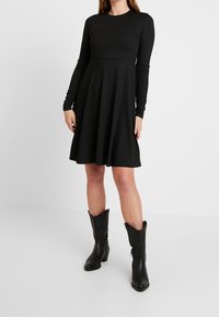 Mustat pitkähihaiset mekko, jossa on ribbitekstuuri, istuva vartalo ja levenevä hame, yhdistettynä polven ylle ulottuviin mustiin nahkasaappaisiin.