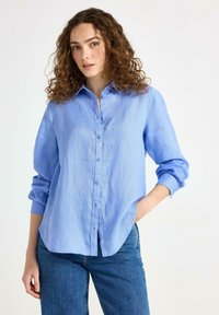 Camicia di lino azzurro chiaro con chiusura a bottoni, maniche lunghe e vestibilità comoda. Presenta un colletto e spacco laterale all'orlo.