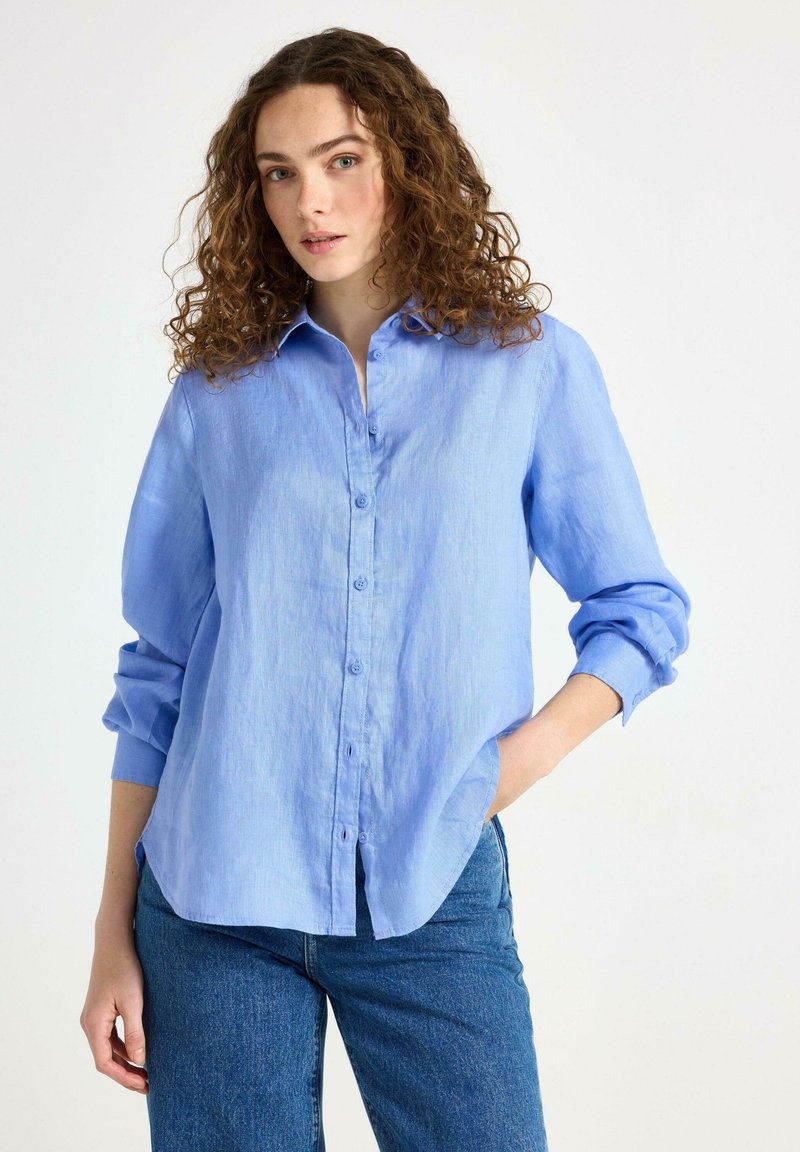 Camicia di lino azzurro chiaro con chiusura a bottoni, maniche lunghe e vestibilità comoda. Presenta un colletto e spacco laterale all'orlo.