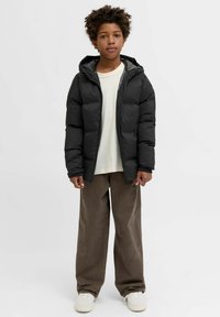 Giacca imbottita nera con cappuccio e zip frontale, abbinata a una camicia chiara e pantaloni larghi di velluto a coste marrone. Scarpe da ginnastica bianche visibili.