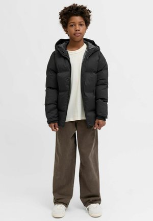 Sort dunjakke med hætte og lynlås foran, kombineret med en lys skjorte og brede brune kordfløjlsbukser. Hvide sneakers er synlige.