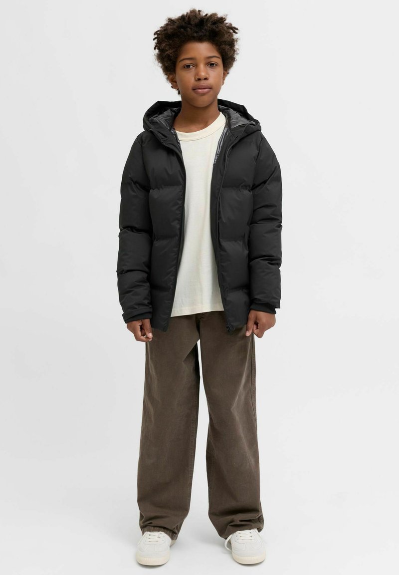 Giacca imbottita nera con cappuccio e zip frontale, abbinata a una camicia chiara e pantaloni larghi di velluto a coste marrone. Scarpe da ginnastica bianche visibili.