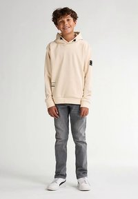 Sudadera con capucha color crema con bolsillo canguro y cordones decorativos, combinada con vaqueros grises y zapatillas blancas.