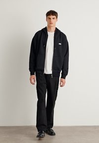 Svart zip-up hettegenser med frontlommer, kombinert med svarte cargos med glidelåsdetaljer. Modellen har på seg en hvit t-skjorte under.