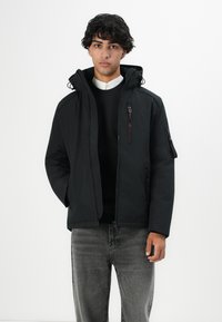 Ung mand iført en sort hættejakke over en sort sweater og en hvid skjorte, parret med slidte grå jeans, står foran en hvid baggrund.
