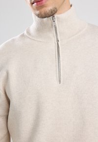 Light beige, zippered high-neck sweater in soft knit fabric. Notable features include ribbed collar and subtle texture.