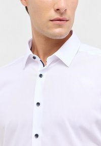 Camicia bianca con colletto aperto, caratterizzata da dettagli dei bottoni blu navy e una texture liscia. Primo piano sulla zona superiore del torso.