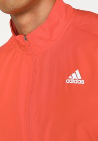 Gros plan d'une personne portant une veste rouge zippée avec un col montant et un logo Adidas blanc sur la poitrine.