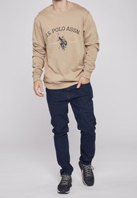 Beige sweatshirt med U.S. Polo Assn. logotyp, rundad halsringning och ribbade ärmslut, parat med mörkblå denimjeans och grå sportskor.