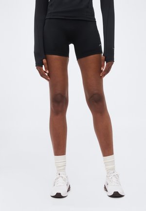 Asmuo dėvi juodus sportinius šortus, ilgomis juodomis rankovėmis marškinėlį, baltas kulkšnies kojines ir baltus sportinius batelius ant paprasto fono.