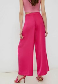 Vrouw in felroze wijde satijnen broek, bijpassende gevederde hoge hakken en een mouwloze roze crop top, staande tegen een witte muur.