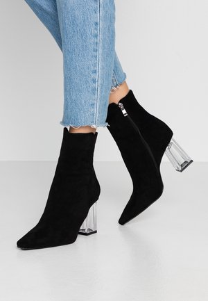 Person trägt hellblaue Jeans und schwarze Wildleder-Stiefeletten mit transparenten Blockabsätzen, steht auf einer glatten weißen Oberfläche.