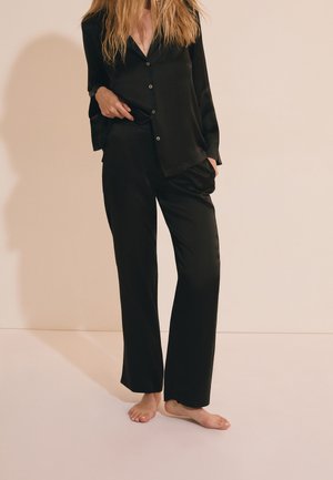 Femme portant un haut noir ample à manches longues boutonné et un pantalon large assorti, debout pieds nus sur fond beige uni.