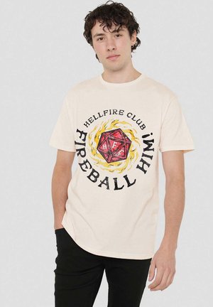 Junger Mann mit lockigem Haar trägt ein cremefarbenes T-Shirt, das einen roten 20-seitigen Würfel zeigt, umgeben von gelben Flammen und dem Text "Hellfire Club Fireball Hm!"