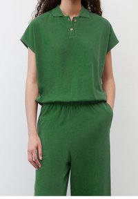 Haut vert à manches courtes avec col polo, fermeture à deux boutons et coupe décontractée. Le matériau semble doux, avec une finition lisse et texturée.