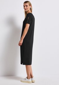 Robe noire longueur genou avec des manches courtes et des fentes sur les côtés. Fabriquée en tissu lisse, elle présente une coupe décontractée et un design simple. Portée avec des baskets blanches.