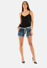 Camiseta de seda negra con tirantes adornados, combinada con pantalones cortos de mezclilla que tienen un cinturón estampado y un lazo. La modelo lleva zapatos de tacón altos negros.
