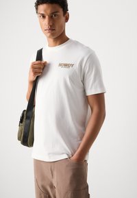 Hvit bomulls t-skjorte med en brun "HOWDY" logo på brystet, kombinert med lysebrune bukser og en grønn skulderveske.