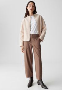 Giacca in pelliccia sintetica beige caldo con zip frontale, abbinata a pantaloni marroni a gamba larga e stivaletti neri a punta. Top bianco semplice sotto.